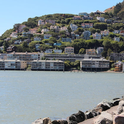 A hillside with numerous houses overlooks a calm body of water, with rocks lining the foreground under a blue sky.