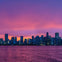 City skyline at dusk with illuminated buildings under a vibrant sunset sky, viewed across a calm body of water.