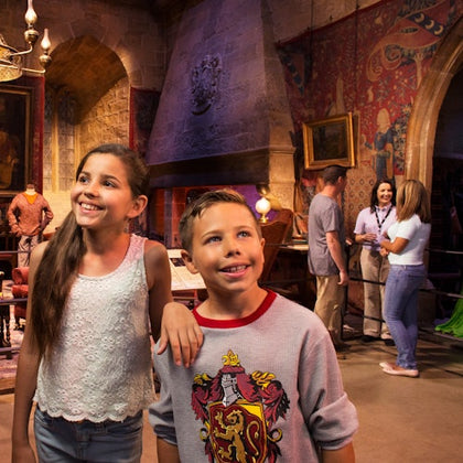 Two children smile inside a room with a medieval theme, featuring paintings, a chandelier, and people in the background.