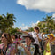 A woman tour guide with a microphone speaks to a group of tourists seated in an open-top bus, surrounded by palm trees and buildings.