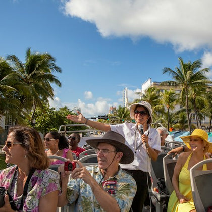 A woman tour guide with a microphone speaks to a group of tourists seated in an open-top bus, surrounded by palm trees and buildings.