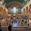 Grand Central Terminal's main concourse with its high arched windows, iconic clock, people walking, and the US flag.