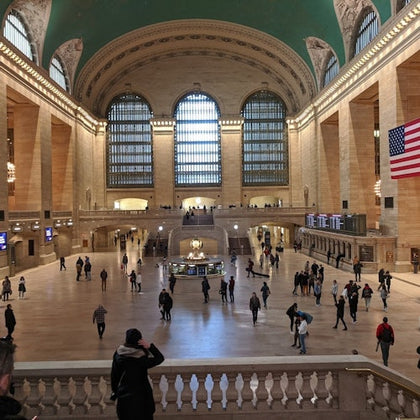 Grand Central Terminal's main concourse with its high arched windows, iconic clock, people walking, and the US flag.