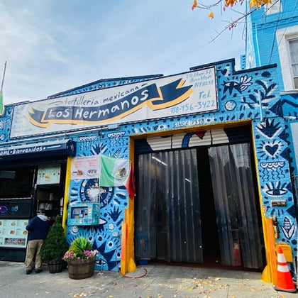 A colorful storefront for Tortilleria Mexicana Los Hermanos, featuring a vibrant mural and a Mexican flag outside.