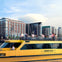 A yellow water taxi with passengers on the top deck in front of a waterfront urban area with modern buildings.