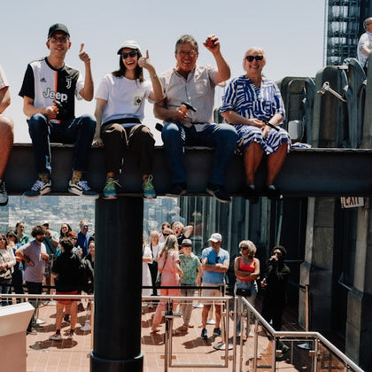 A group of people sitting on a railing above a crowd on a sunny day, with an urban cityscape in the background.