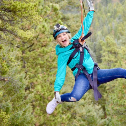 A person wearing a helmet and harness is ziplining through a forested area, smiling enthusiastically.