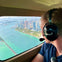 Person wearing a headset looks out of a small airplane window at a city with tall buildings and a coastline along turquoise waters.