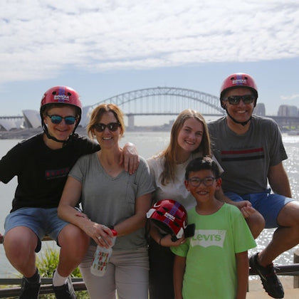 Group of five people smiling, posing near water with a bridge and cityscape in the background, some wearing helmets.