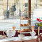A table set with tea, pastries on a tiered stand, and champagne glasses by a window overlooking a street scene.