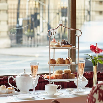 A table set with tea, pastries on a tiered stand, and champagne glasses by a window overlooking a street scene.