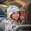 A young child in a patterned jacket smiles while sitting on a bus, with another person seated nearby.