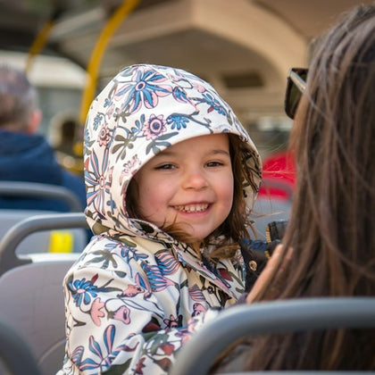 A young child in a patterned jacket smiles while sitting on a bus, with another person seated nearby.