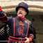 A beefeater in a traditional black and red uniform stands in front of a stone archway, raising one hand while speaking.