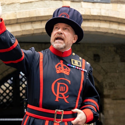 A beefeater in a traditional black and red uniform stands in front of a stone archway, raising one hand while speaking.