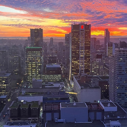 Cityscape at dusk with illuminated buildings and a vibrant orange and purple sunset in the background.