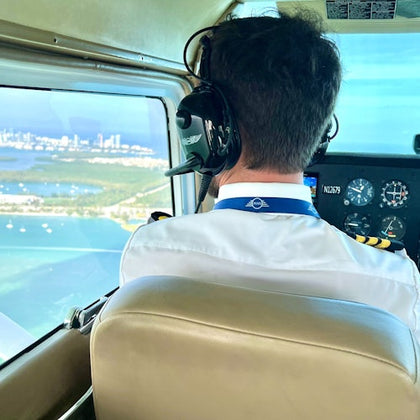Pilot wearing headphones flying a plane with a view of the coastline and cityscape visible through the cockpit window.