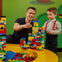 An adult and a child building colorful structures with plastic blocks on a yellow table in a playroom.