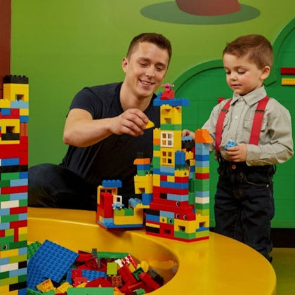 An adult and a child building colorful structures with plastic blocks on a yellow table in a playroom.