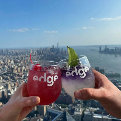 Two hands holding drinks with "edge New York" logos, overlooking a cityscape and water under a clear sky.