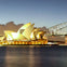 The Sydney Opera House illuminated at night with the Sydney Harbour Bridge in the background, viewed from across the water.