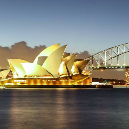 The Sydney Opera House illuminated at night with the Sydney Harbour Bridge in the background, viewed from across the water.