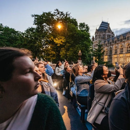 A group of people on an open-top bus tour observing and taking photos near a historic building at dusk.