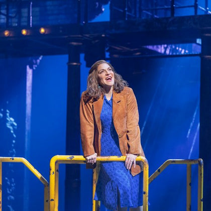 A woman in a blue dress and brown jacket stands behind a yellow railing on a stage with a blue-lit industrial setting.