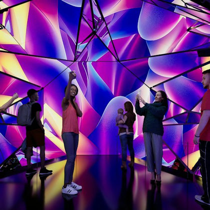 People standing in a vibrant, mirrored room with colorful abstract lighting and reflections, some taking photos.