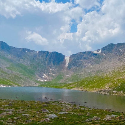 A serene mountain lake surrounded by rocky terrain and green grass under a partly cloudy sky.