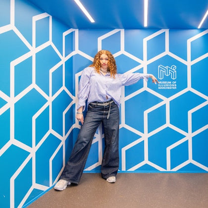 A person stands in a blue room with geometric wall patterns and a sign reading 'Museum of Illusions Manchester.'