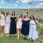 A group of six women posing outdoors in a vineyard on a sunny day, dressed in casual summer dresses, with a scenic background.