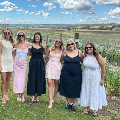 A group of six women posing outdoors in a vineyard on a sunny day, dressed in casual summer dresses, with a scenic background.