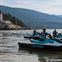 Several people on jet skis in calm water near a lighthouse on a rocky shoreline, with forested mountains in the background.