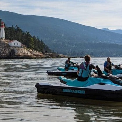 Several people on jet skis in calm water near a lighthouse on a rocky shoreline, with forested mountains in the background.