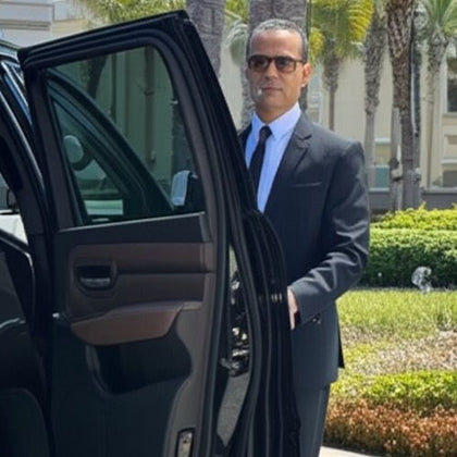 A man in a dark suit and sunglasses stands beside an open car door in a sunny, landscaped area with a modern building in the background.