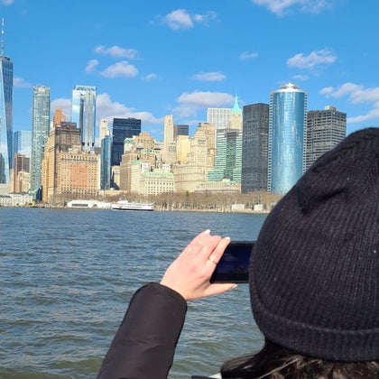 A person in a black hat takes a photo of a city skyline across the water on a clear day with blue skies.