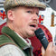 A man wearing a flat cap speaks into a microphone, with British-themed decorations visible in the background.