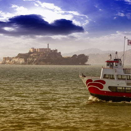A ferry with red and white colors in the foreground and an island with a prominent building in the background, under a cloudy sky.