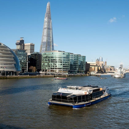 A boat navigates the River Thames with modern buildings, including The Shard, visible against a blue sky.