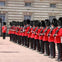A row of guards in red uniforms and black bearskin hats stand in formation outside a stone building with multiple windows.