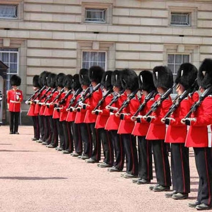 A row of guards in red uniforms and black bearskin hats stand in formation outside a stone building with multiple windows.