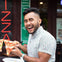 A person smiling while holding a slice of pizza, sitting at an outdoor table near a restaurant with a neon pizza sign.