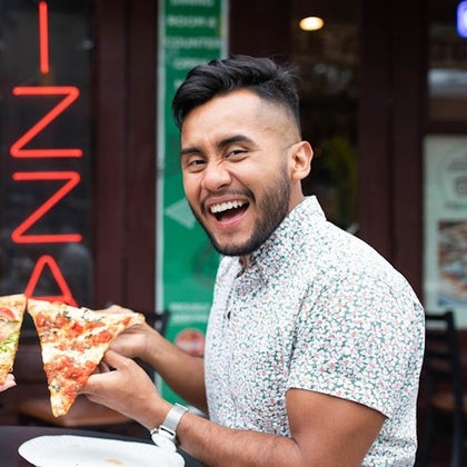 A person smiling while holding a slice of pizza, sitting at an outdoor table near a restaurant with a neon pizza sign.