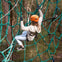 Person wearing an orange helmet and harness climbing a green rope net in a forested area.