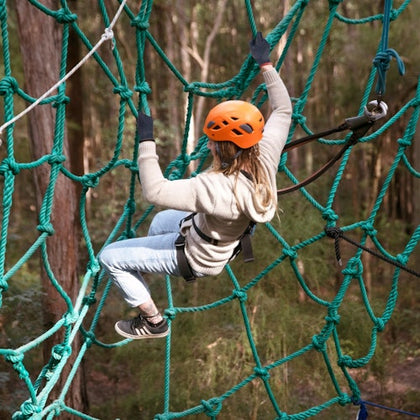 Person wearing an orange helmet and harness climbing a green rope net in a forested area.