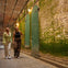 Two people walking in a covered walkway with green moss-covered walls and decorative doors, illuminated by warm lights.