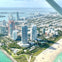 Aerial view of a coastal city with tall buildings, green parks, and a sandy beach with people sunbathing and swimming.