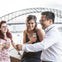 A group of people smiling and holding drinks near a bridge on a cloudy day.