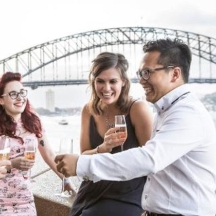 A group of people smiling and holding drinks near a bridge on a cloudy day.